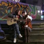 Two people sharing guitar on steps, graffiti