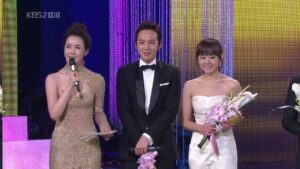 Three presenters on stage holding flowers and microphone