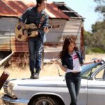Man playing guitar on car hood, woman nearby