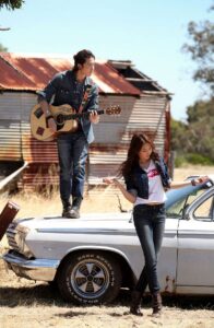 Man playing guitar on car hood, woman nearby