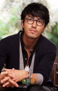Seated man with glasses, necklace, and cardigan