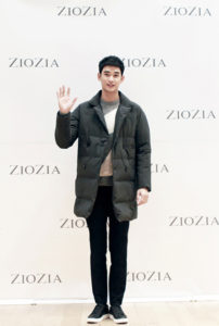 Person in coat waving at event backdrop.