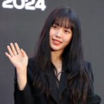 Woman waving, wearing a black outfit, smiling.