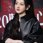 Woman in black outfit, seated, serious expression.