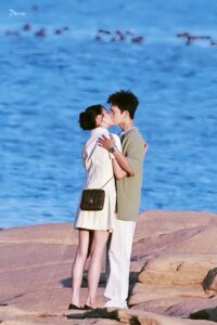 Couple kissing on rocky shore by ocean.
