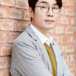 Man in glasses leaning against brick wall.