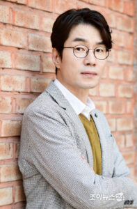 Man in glasses leaning against brick wall.